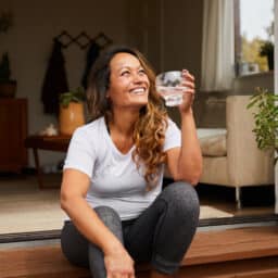 Smiling woman drinking a glass of water sitting on the ledge of a door that opens to a yard, her living room can be seen behind her.